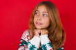 © Jihan - Happy little kid girl wearing knitted sweater christmas over red background anticipates something awesome happen, looks happily aside, keeps hands together near face, has glad expression.