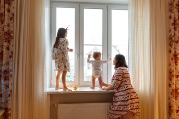 Family Christmas photo. Mom and two daughters are playing on the windowsill. It's winter outside and snow is falling