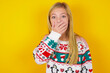 © Roquillo - Emotional caucasian little kid girl wearing knitted sweater christmas over yellow background gasps from astonishment, covers opened mouth with palm, looks shocked at camera. Reaction concept