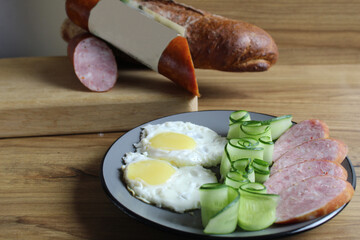 Canvas Print - breakfast traditional scrambled eggs fried eggs two ham sausages and cucumber salad on a gray plate on the table. next to it are products for cooking breakfast sausage bread. Horizontal photo