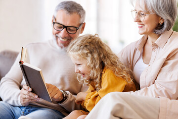 Cute little girl granddaughter smiling when reading book with senior grandparents