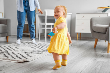 Wall Mural - Cute baby girl in yellow dress learning to walk with mother at home