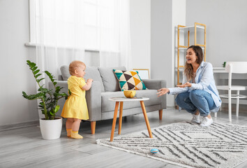 Wall Mural - Cute baby girl in yellow dress learning to walk with mother near sofa at home