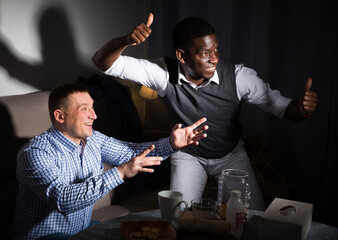 Two excited male interracial friends watching football game at home together