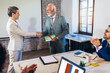 © Mediteraneo - Senior businessman receiving award from businesswoman in front of business professionals, applauding at business seminar in office building