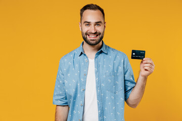 Wall Mural - Young satisfied rich cool smiling happy caucasian man 20s wearing blue shirt white t-shirt hold in hand credit bank card isolated on plain yellow background studio portrait. People lifestyle concept.