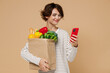 © ViDi Studio - Young smiling fun vegetarian woman in casual clothes hold paper bag with vegetables hold in hand use mobile cell phone read browsing menu isolated on plain pastel beige background Shopping concept.