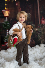 Sticker - Cute fashion toddler boy, playing in the snow with teddy bear in front of a wooden cabin log