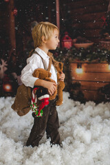 Sticker - Cute fashion toddler boy, playing in the snow with teddy bear in front of a wooden cabin log