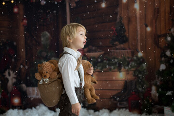 Wall Mural - Cute fashion toddler boy, playing in the snow with teddy bear in front of a wooden cabin log