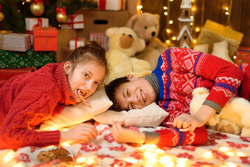 Wall Mural - Portrait of boy and girl in new year or christmas decoration. They lie and eat sweets and cookies. Holiday lights, gifts and christmas tree decorated with toys.