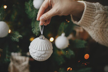Hand In Cozy Sweater Holding Modern White Bauble At Christmas Tree Lights. Woman Decorating Stylish Boho Christmas Tree With Vintage Ornament. Winter Holidays Preparations. Atmospheric Hygge Home