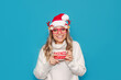 © Марина Демешко - A young cute smiling blonde woman in glasses with deer antlers, a white warm sweater, Christmas Santa hat,red mittens holds big deer mug with hot drink in her hands isolated on color blue background