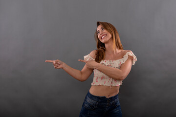 Cheerful brunette model dressed in casual clothes smiles and points sideways with finger isolated on gray background