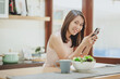 © interstid - Happy smiling Asian woman using smartphone mobile at home in kitchen with salad bowl on the table