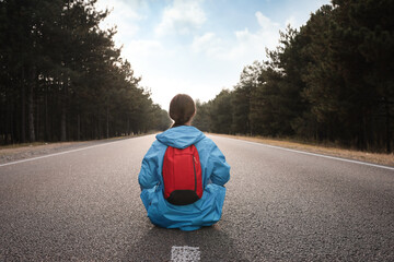 Poster - Young woman with backpack sitting on road near forest, back view