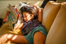 A Child Lays On Couch In Dappled Window Light Reading Chapter Book