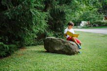 A Child Sits On Boulder By Side Of Road Reading Picture Book