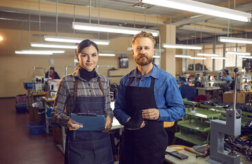 Portrait of two happy young shoe factory workers in aprons standing in big workshop, holding new leather boot and clipboard and looking at camera. Concept of modern footwear manufacturing industry