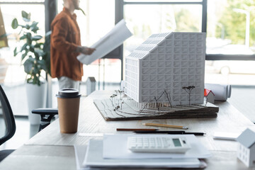 Wall Mural - selective focus of house model and paper cup on desk near architect standing with paper on blurred background