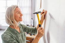 Happy Smiling Middle Aged Woman Hammering Nail To Wall