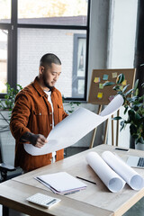 Sticker - asian engineer looking at blueprint while standing near papers and gadgets on work desk