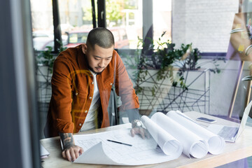 Wall Mural - asian architect looking at construction plan while standing at workplace on blurred foreground