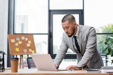 bearded asian businessman standing near work desk and looking at laptop in office