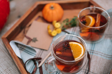 Close up transparent cups with hot spiced tea with star anise, cinnamon stick and a slice of orange on the wooden tray with decorative pumpkin, flowers and blank postcard. Cozy autumn. Tea time ideas.