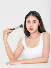 Wall Mural - Portrait beauty studio shot of millennial Asian short black hair female model in tank top vest undershirt place arm on table holding concealer brush in hand look at camera on white background