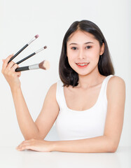 Wall Mural - Portrait beauty studio shot of Asian short black hair female model with teeth retainer in tank top vest undershirt hold three size foundation brushes in hand smile look at camera on white background