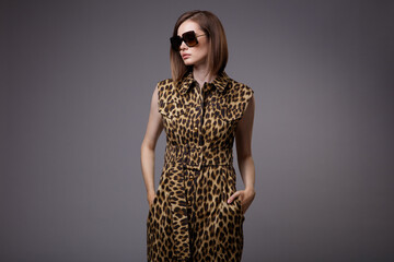 High fashion photo of a beautiful elegant young woman in a pretty shirt dress sleeveless animal leopard print, black ankle boots, stylish sunglasses posing over gray background. Slim figure.