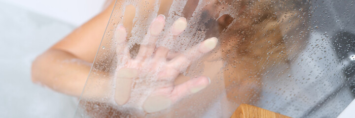 Naked woman sitting in bathtub and holding her hand on misted glass closeup