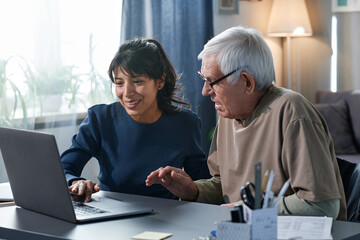 friendly volunteer teaching to use the computer to senior man while they sitting at the table at hom
