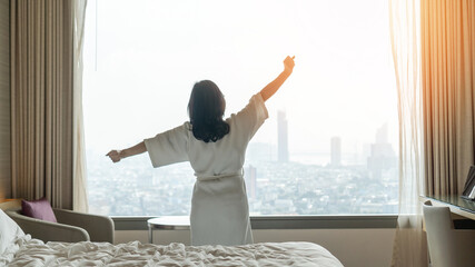 Wall Mural - Hotel relaxation on lazy day with Asian woman waking up from good sleep on bed in weekend morning resting in comfort bedroom looking toward city view, having happy, work-life quality balance lifestyle