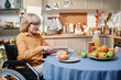 Senior woman using wheelchair sitting at the table and cutting bread, she cooking breakfast for herself in the kitchen