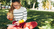 Horizontal shot of a student female in casual outfit, eyeglasses, sitting on the green grass at the college campus, have lunch, and studying. Woman takes a rest eating fast food and learning outside.