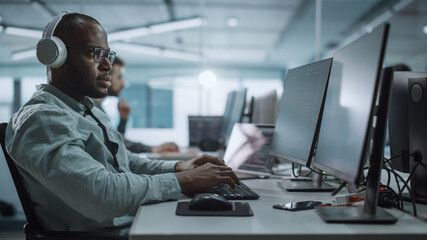 Office: Energetic Black IT Programmer Puts on Headphones Starts Working on Desktop Computer. Male Website Developer, Software Engineer Developing App, Video Game. Listening to Podcast, Music