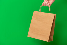 Female Hand Holding Brown Craft Blank Shopping Bag Isolated On Green Background. Black Friday Sale, Discount, Recycling, Shopping And Ecology Concept.