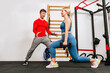 Young blond woman doing weighted lunge with dumbbells, with personal trainer motivating her. Male coach helping sportswoman to do exercises with dumbbell at gym. Sports and healthcare concept.