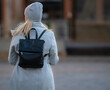 Happy young blonde teenage girl going to school or college wearing black backpack. Blonde girl in rgay hat and gray coat. Pictuce from back