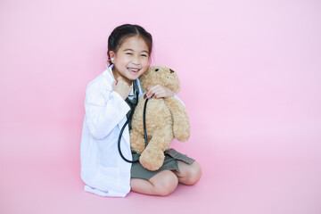 Wall Mural - Little asian child girl dreaming about is a veterinarian using stethoscope with teddy bear on pink background