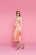 © LIGHTFIELD STUDIOS - Smiling woman in silk dress and sunglasses walking on pink background