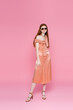 © LIGHTFIELD STUDIOS - Full length of smiling woman in dress and sunglasses posing on pink background