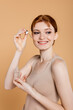 © LIGHTFIELD STUDIOS - Smiling redhead woman in top applying cosmetic serum isolated on beige