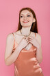 © LIGHTFIELD STUDIOS - Smiling redhead woman in peach dress touching necklace isolated on pink