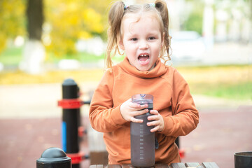 A little girl walks on the city's playground in the warm season and drinks water from a shaker. Lifestyle sport