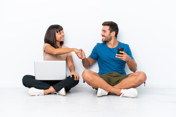 Wall Mural - Young couple sitting on the floor holding pc and mobile phone isolated on white background handshaking after good deal