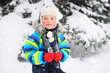 © Evgeniy Kalinovskiy - a small cute baby boy in a striped winter jacket, hat and red mittens stands looking at the camera against the background of snowfall and forest.