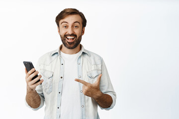 Cheerful bearded man pointing at cellphone and smiling amazed, found smth online, standing over white background